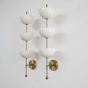May include: Two mid-century modern wall sconces. Each sconce features three white, bowl-shaped shades stacked vertically on a brass rod. The sconces have brass mounting plates and small brass finials. The design is minimalist and elegant.