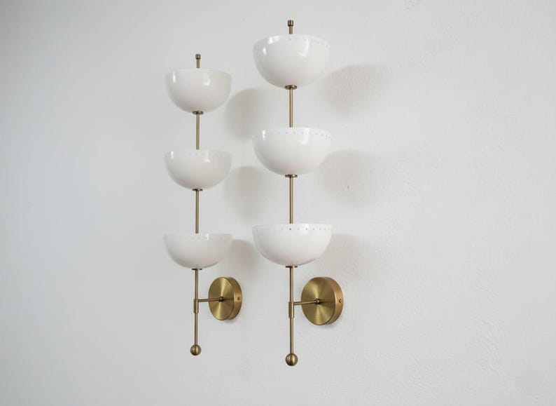 May include: Two mid-century modern wall sconces. Each sconce features three white, bowl-shaped shades stacked vertically on a brass rod. The sconces have brass mounting plates and small brass finials. The design is minimalist and elegant.