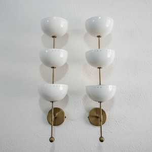 May include: Two matching wall sconces with a mid-century modern design. Each sconce features three white, bowl-shaped shades stacked vertically, connected by brass-colored stems and mounted on a brass-colored circular base.
