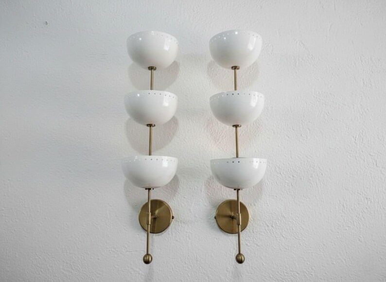 May include: Two matching wall sconces with a mid-century modern design. Each sconce features three white, bowl-shaped shades stacked vertically, connected by brass-colored stems and mounted on a brass-colored circular base.
