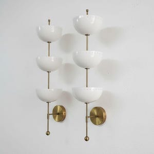 May include: Two mid-century modern wall sconces. Each sconce features three white, bowl-shaped shades stacked vertically on a brass rod. The sconces have brass wall mounts and are mounted on a white wall.