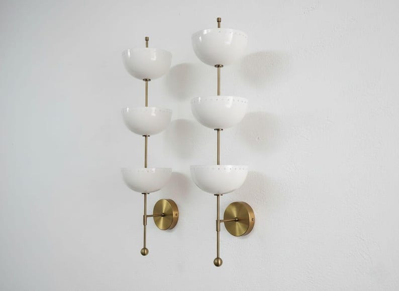 May include: Two mid-century modern wall sconces. Each sconce features three white, bowl-shaped shades stacked vertically on a brass rod. The sconces have brass wall mounts and are mounted on a white wall.