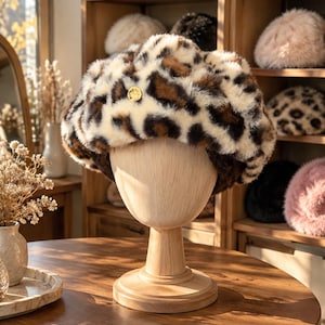 May include: A fluffy faux fur beret featuring a brown, black, and cream leopard print pattern. The hat is displayed on a wooden mannequin head and includes a small, circular gold-toned metal emblem centered on the front of the plush fabric.