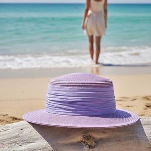 May include: A lilac sun hat with a wide brim and a fabric band rests on driftwood on a sandy beach. The hat is in focus, with the sea and a person in the background.