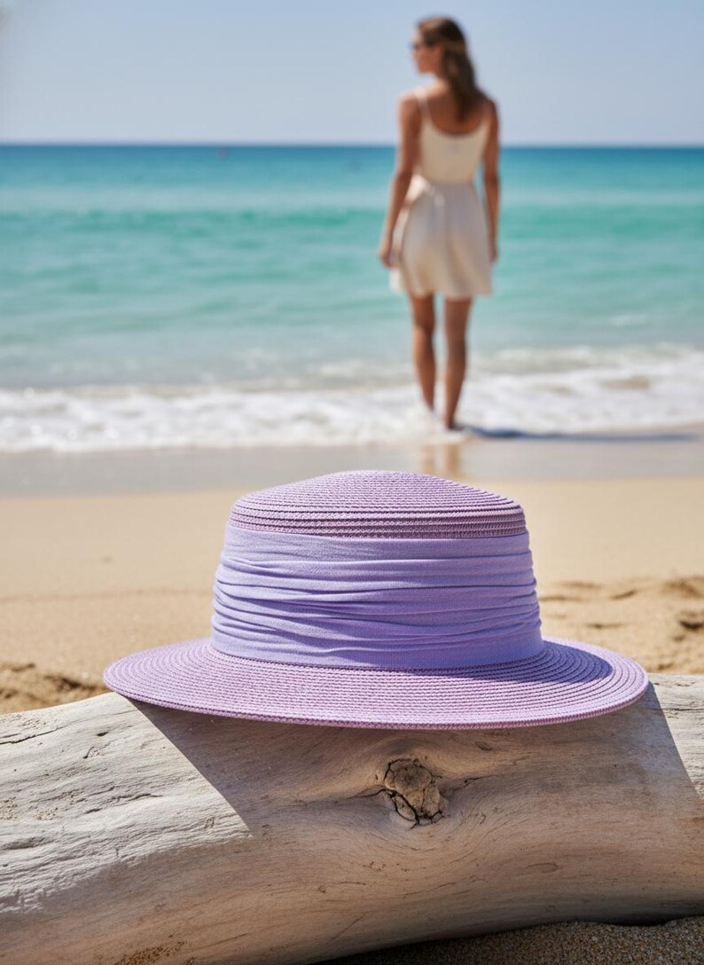 May include: A lilac sun hat with a wide brim and a fabric band rests on driftwood on a sandy beach. The hat is in focus, with the sea and a person in the background.