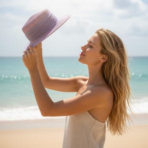May include: A lilac sun hat with a wide brim is held up against a sunny sky. The hat is the focal point, with a beach and ocean in the background. The hat appears to be made of woven material.