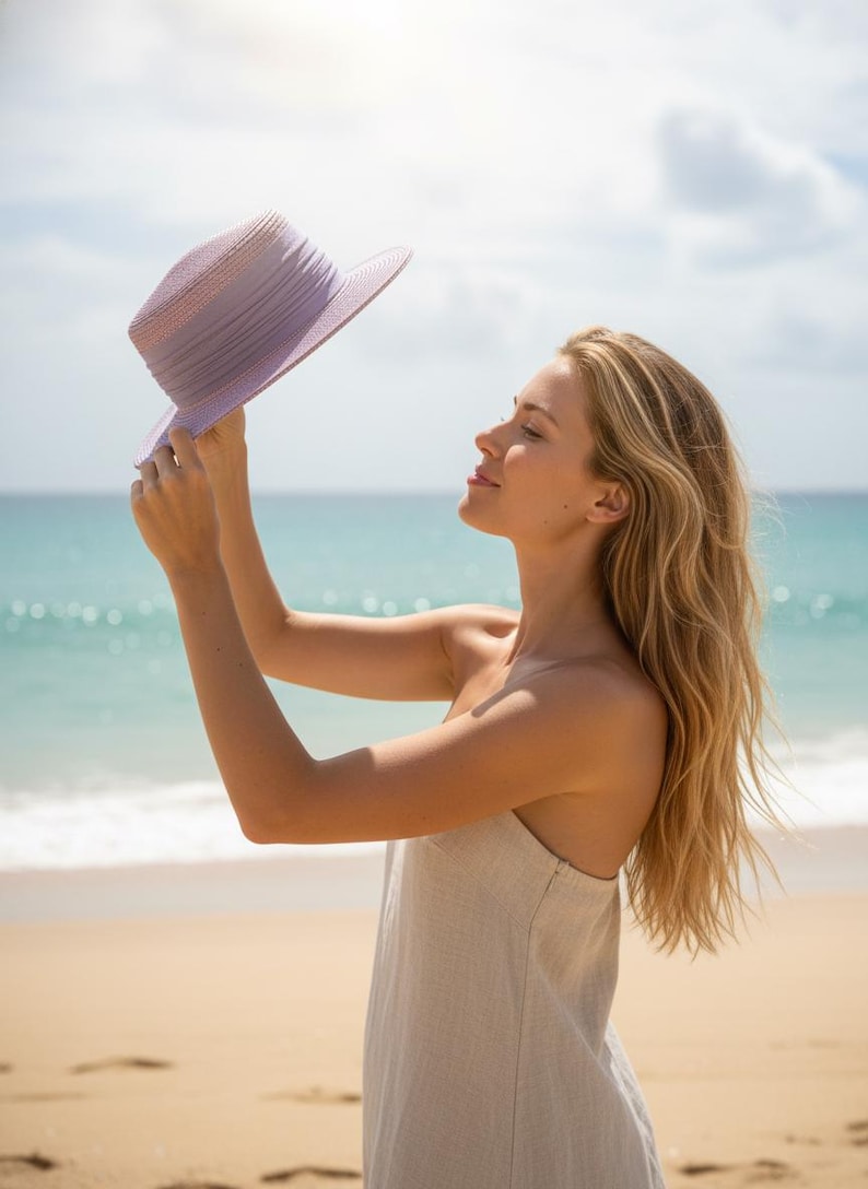 May include: A lilac sun hat with a wide brim is held up against a sunny sky. The hat is the focal point, with a beach and ocean in the background. The hat appears to be made of woven material.