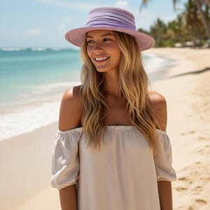 May include: A woman wearing a lavender hat with a wide brim and a light beige off-the-shoulder dress. The background shows a beach with turquoise water and palm trees, creating a summery, relaxed vibe.