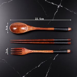 May include: A wooden spoon (22.5 cm long, 4 cm wide), chopsticks, and a fork (3 cm wide) set. The utensils are brown with black leather-wrapped handles and a dark wood finish. The set is displayed on a black marble surface.