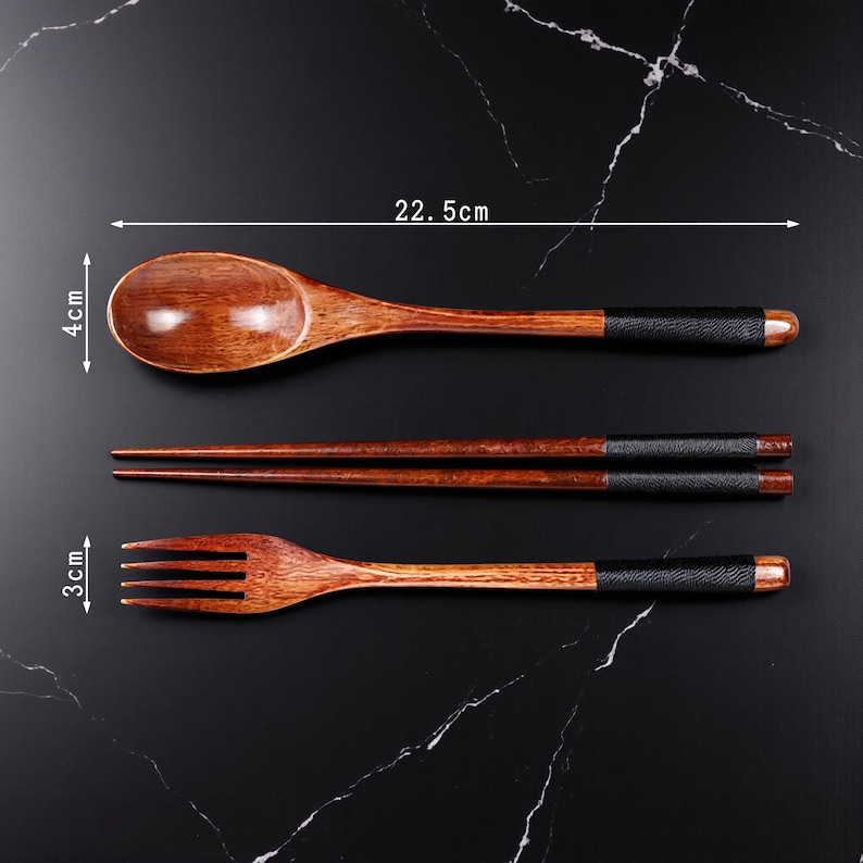 May include: A wooden spoon (22.5 cm long, 4 cm wide), chopsticks, and a fork (3 cm wide) set. The utensils are brown with black leather-wrapped handles and a dark wood finish. The set is displayed on a black marble surface.