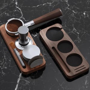 May include: Coffee accessories set on a black marble surface. The set includes a portafilter, tamper, brush, and wooden stands. The portafilter has a wooden handle and a metal basket. The tamper is silver. The wooden stands have circular cutouts.