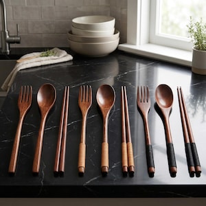 May include: Wooden cutlery set featuring forks, spoons, and chopsticks. The spoons and chopsticks have a dark brown finish, while the forks have a lighter brown finish. Some chopsticks and spoons have textured grips. The set is arranged on a black countertop.