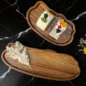 Puede incluir: Dos bandejas de madera con un borde ondulado único. Una bandeja contiene dos rebanadas de pastel con coloridas coberturas. La otra bandeja está vacía, con una ramita de flores blancas secas. Las bandejas están sobre una superficie de mármol negro.