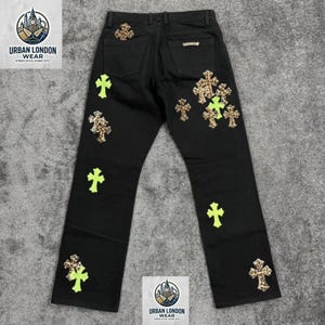 Puede incluir: Vaqueros negros con motivos de cruces en estampado de leopardo y verde neón. Los vaqueros tienen un diseño clásico de cinco bolsillos y corte recto. El logotipo de la marca "URBAN LONDON WEAR" es visible.