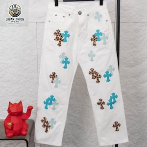 May include: White denim jeans adorned with cross-shaped patches in leopard print, light blue, and pale blue. The jeans have a classic five-pocket design and a straight leg fit. The Urban London Wear logo is visible in the upper left corner.