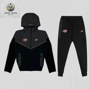 Può includere: Completo tuta nero con giacca con cappuccio e pantaloni abbinati. La giacca ha una chiusura a zip, tasche laterali e un logo sul petto. I pantaloni hanno una vestibilità slim e un logo sulla gamba sinistra. Di Urban London Wear.