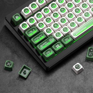 Könnte beinhalten: Eine mechanische Tastatur mit weißen und grünen Tastenkappen. Die Tastatur hat einen schwarzen Rahmen und grüne LED-Beleuchtung. Die Tastenkappen sind eine Mischung aus Weiß mit grüner Beschriftung und Grün mit weißer Beschriftung. Mehrere lose Tastenkappen liegen herum.