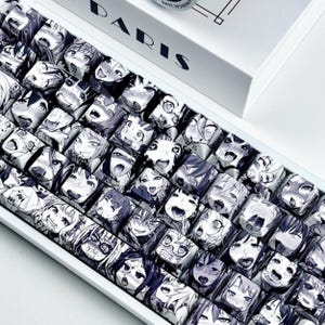 Puede incluir: Un teclado mecánico blanco con teclas personalizadas con caras de personajes de estilo anime en blanco y negro. El teclado está sobre un fondo blanco, con una caja que dice "PARIS" en letras negras.