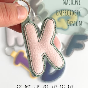 May include: A pink letter "K" keyring with a green border and silver ring. The keyring is held in a hand, with other letter keyrings in the background. Text on the image reads "Machine Embroidery Design" and file types .PES .DST .HUS .VP3 .XXX .JEF .EXP.