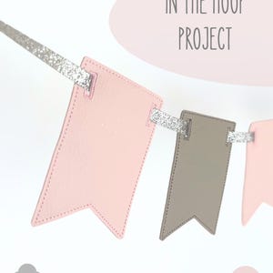 May include: A bunting banner featuring alternating light pink and taupe faux leather pennants suspended on a silver glitter ribbon. The text IN THE HOOP PROJECT is displayed at the top, and the file formats .PES .VP3 .XXX .HUS .EXP .JEF .DST are listed below.