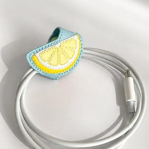 May include: A light blue felt cable organizer shaped like a lemon slice, with yellow and white details, encircling a white charging cable. The cable is coiled, and the organizer is stitched with yellow thread.