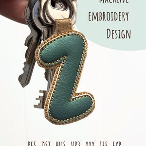 Peut inclure: Un porte-clés en cuir vert d'eau en forme de chiffre 1, avec des coutures dorées et un dos en cuir beige. Le porte-clés est attaché à un jeu de clés. Le texte sur l'image indique "MACHINE EMBROIDERY DESIGN" et les types de fichiers .PES .DST .HUS .VP3 .XXX .JEF .EXP.