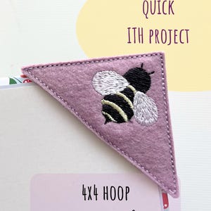 Könnte beinhalten: Ein lavendelfarbener Eck-Lesezeichen aus Filz mit einem gestickten Bienen-Design. Die Biene hat schwarz-gelbe Streifen, weiße Flügel und schwarze Details. Der Text "QUICK ITH PROJECT" befindet sich in einer gelben Blase. Der Text "4x4 HOOP" steht in einem weißen Rechteck.