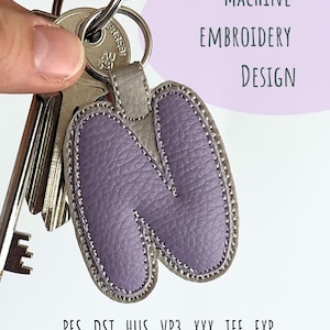 May include: A key ring with a lavender letter "N" design. The letter is stitched with a silver thread and has a gray leather backing. The key ring is attached to a set of keys. The text "MACHINE EMBROIDERY DESIGN" is in the background.