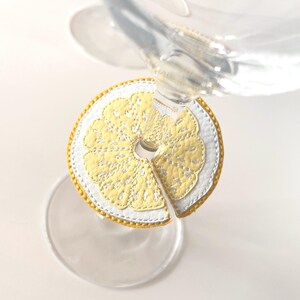Pode incluir: Uma base de copo decorativa em forma de fatia de limão, feita de couro amarelo e branco com uma borda dourada. A base de copo foi projetada para caber em volta da haste de uma taça de vinho. Close-up em fundo branco.