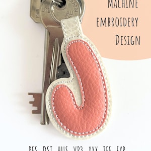 Può includere: Un portachiavi con chiavi e un ciondolo con l'iniziale "J" in similpelle. Il ciondolo è rosa corallo con un bordo bianco e un contorno cucito bianco. Il testo "MACHINE EMBROIDERY DESIGN" è su uno sfondo color pesca.