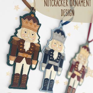Può includere: Tre ornamenti schiaccianoci ricamati con nastri, che includono il testo ITH NUTCRACKER ORNAMENT DESIGN e i formati di file .PES .DST .JEF .HUS .XXX .VP3 .EXP. Gli schiaccianoci sono realizzati in marrone, blu e rosso con cuciture dettagliate.