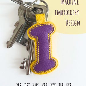 May include: A purple letter "I" keychain with a yellow border, attached to a keyring with keys. The keychain has a stitched design. Text on the image reads "MACHINE EMBROIDERY DESIGN".