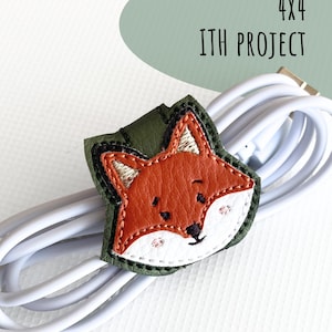May include: A fox-shaped cable organizer made of olive green faux leather with an embroidered orange and white fox face. The organizer is wrapped around a white charging cable. Text on the image reads 4x4 ITH PROJECT and .PES .DST .JEF .HUS .XXX .VP3 .EXP.