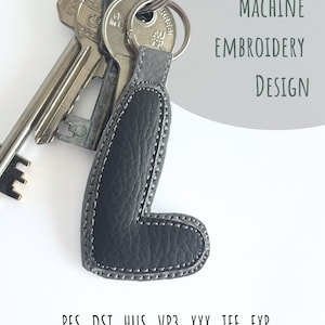 May include: A set of keys with a gray leather keychain in the shape of the letter "L". The keychain is stitched with white thread and has the words "Machine Embroidery Design" in a light green oval. File formats are listed below the keychain.