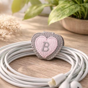 May include: A gray heart-shaped cable organizer with a pink heart in the center, embroidered with the letter "B". The organizer is resting on a white cable, with the earbuds visible. The background includes a wooden surface and a potted plant.