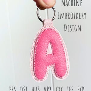 May include: A pink leather keychain in the shape of the letter "A", with a light pink border and a silver key ring. The text "MACHINE EMBROIDERY DESIGN" is visible. File formats: .PES .DST .HUS .VP3 .XXX .JEF .EXP.