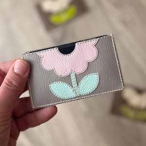 Puede incluir: Tarjetero de cuero gris con un diseño de flor rosa y hojas verde menta. La flor y las hojas están cosidas en el tarjetero. Los bordes están ribeteados con costuras doradas. El tarjetero se sostiene en una mano.