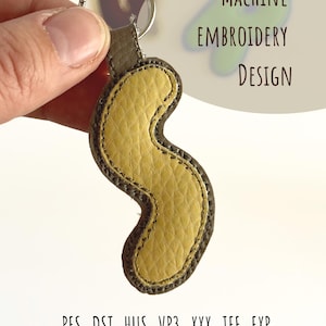 May include: A yellow and brown leather keychain with a wavy design. The keychain is attached to a silver ring. The text "MACHINE EMBROIDERY DESIGN" is visible on the right side of the image. Below the keychain are the file extensions .PES .DST .HUS .VP3 .XXX .JEF .EXP.