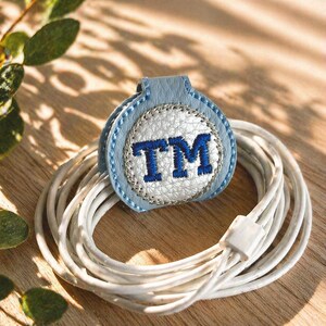 以下が含まれることがあります： 紺色の「TM」の刺繍が入った、青い革製のケーブルオーガナイザー。 オーガナイザーは銀色の糸で縫い付けられており、白い充電ケーブルと組み合わせています。 背景は明るい茶色の木製の表面です。