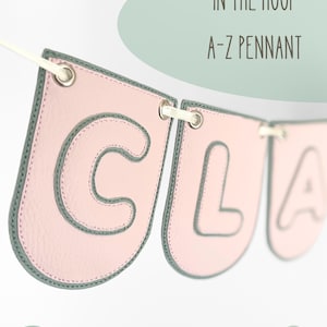 Könnte beinhalten: Eine Reihe hellrosa Wimpel mit dunkelgrauen gestickten Rändern und dunkelgrauen Buchstaben, darunter C und L, auf einem weißen Band. Der Text IN THE HOOP A-Z PENNANT sowie die Dateiformate PES, DST, JEF, HUS, XXX, VP3, EXP sind zu sehen.