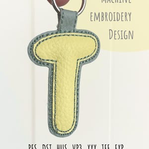 May include: A yellow leather-like letter "T" keychain with a gray border and a silver keyring. The keychain is held up by a hand. The text "MACHINE EMBROIDERY DESIGN" is visible. Below the keychain are embroidery file formats.