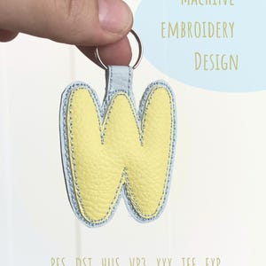May include: A yellow leather keychain shaped like the letter "W", with a light blue border and stitching. It's attached to a silver ring and a light blue leather strap. The image includes the text "MACHINE EMBROIDERY DESIGN".