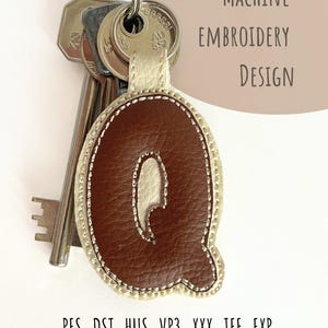 May include: A brown leather keychain with the letter "Q" stitched in the center, attached to a silver keyring with keys. The keychain has a cream-colored border and the text "MACHINE EMBROIDERY DESIGN" is visible. File formats .PES .DST .HUS .VP3 .XXX .JEF .EXP are listed.