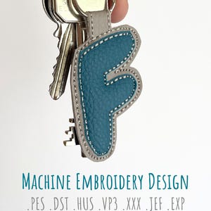 May include: Teal letter "F" keyring with a grey border and white stitching, attached to a silver keyring with keys. The keyring is made of textured leather. Text at the bottom reads "MACHINE EMBROIDERY DESIGN".