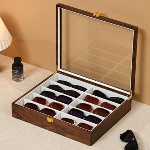 May include: A rectangular wooden storage box with a clear glass lid and a gold-colored metal latch. The interior is divided into twelve white-lined compartments, each containing a pair of sunglasses in various colors including black and brown.