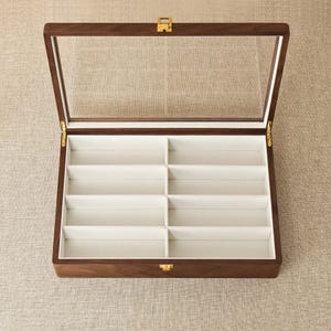 May include: A rectangular wooden display case with a hinged glass lid and a gold-colored metal latch. The interior features eight individual compartments lined with a light-colored fabric, designed for organizing items in a structured grid layout.