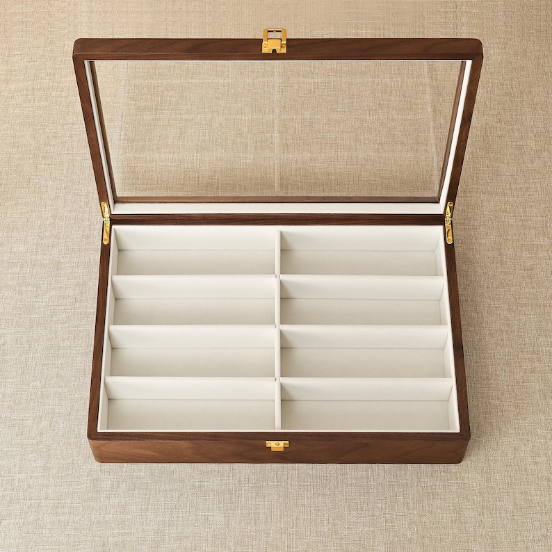 May include: A rectangular wooden display case with a hinged glass lid and a gold-colored metal latch. The interior features eight individual compartments lined with a light-colored fabric, designed for organizing items in a structured grid layout.