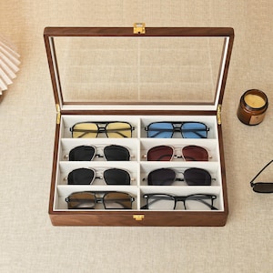 May include: A rectangular wooden display case with a hinged glass lid and a gold-tone latch, featuring eight individual white-lined compartments. Inside are eight pairs of sunglasses with various frame styles and lens colors, including yellow, blue, black, and brown.