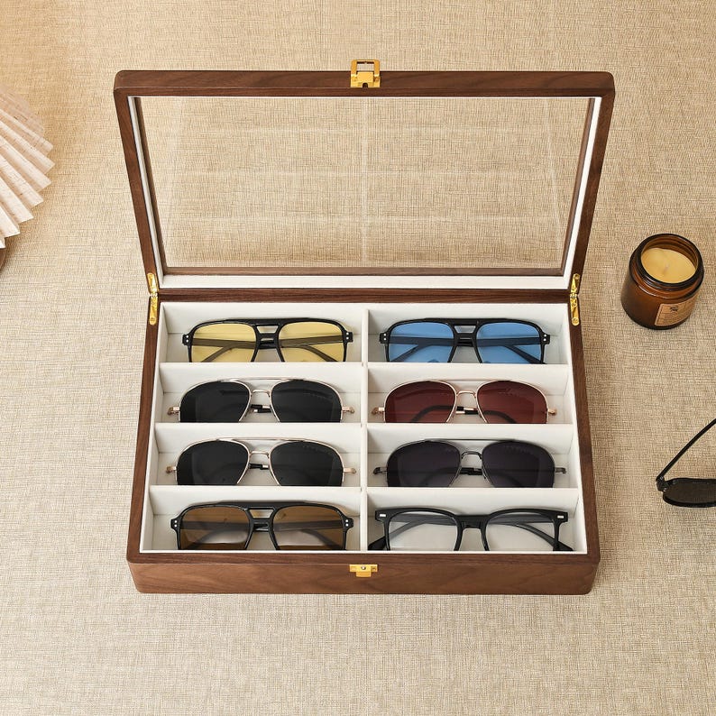 May include: A rectangular wooden display case with a hinged glass lid and a gold-tone latch, featuring eight individual white-lined compartments. Inside are eight pairs of sunglasses with various frame styles and lens colors, including yellow, blue, black, and brown.