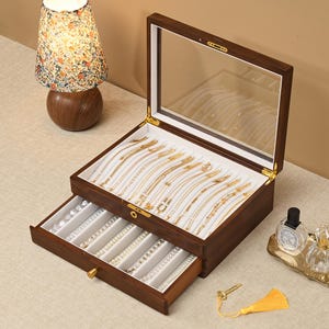 May include: A dark wood jewelry box with a glass lid and a pull-out drawer. The interior is lined with white velvet and features multiple slots for organizing necklaces and bracelets. A small gold-colored key with a yellow tassel is included.